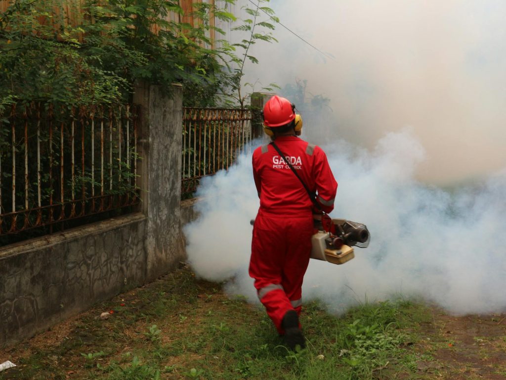 A worker in protective gear applies termite treatment around a building foundation.